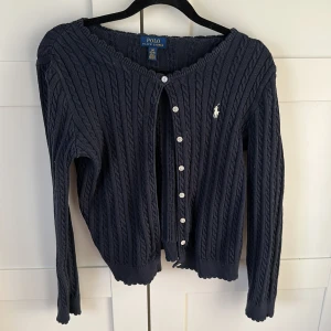 Marinblå kofta från Ralph Lauren - Säljer en stilren marinblå kofta från Ralph Lauren med knappar framtill och det klassiska logotypbroderiet på bröstet. Koftan har långa ärmar och en ribbad struktur som ger en elegant touch. Perfekt för att hålla sig varm och snygg. Tröjan är storlek 12-14 år så ungefär xs/xxs.      Koftan är i mycket gott skick och endast andvänd ett fåtal gånger