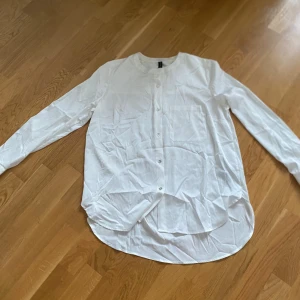 Vit långärmad skjorta - Säljer en stilren vit långärmad skjorta med knappar framtill. Skjortan har en klassisk design med rundad nederkant och är perfekt för en minimalistisk look. Har en liten smink fläck vid kragen 
