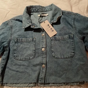 Blå jeansjacka från NA-KD - Snygg blå jeansjacka från NA-KD med en croppad design och fransig nederkant. Jackan är helt helt oandvänd och prislappen sitter fortfarande kvar. Säljer då den inte passar🙏Perfekt för en avslappnad stil💗