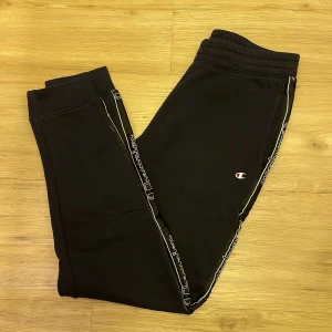 Svarta mjukisbyxor från Champion - Säljer ett par svarta mjukisbyxor från Champion med elastisk midja och logotyp längs sidorna. Perfekta för en avslappnad stil. Byxorna har en bekväm passform och är gjorda i mjukt material.