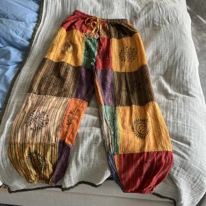 Färgglada bomullsbyxor med mönster - Säljer ett par färgglada byxor i 100% bomull, tillverkade i Nepal. Byxorna har ett patchwork-mönster i olika färger som röd, grön, orange och blå. De har en elastisk midja med snörning och unika tryckdetaljer.