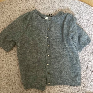 Grå kofta med korta ärmar - Säljer en stilren grå kofta med korta ärmar och knappar framtill. Perfekt för lager-på-lager och passar till många olika outfits. Koftan har en mjuk och bekväm känsla.