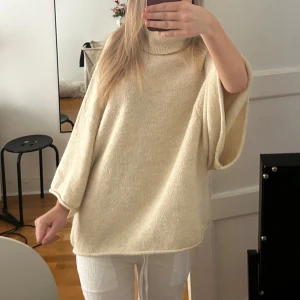 Beige stickad poncho - Säljer en mysig beige stickad poncho med polokrage. Perfekt för kyliga dagar och ger en avslappnad look. Den har vida ärmar och en lös passform som gör den bekväm att bära. Passar bra till jeans eller leggings. Slutsåld på hemsidan.