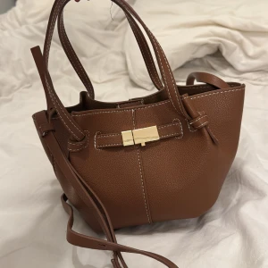 Brun axelväska med gulddetalj - Snygg brun axelväska i skinn med en elegant gulddetalj framtill. Väskan har justerbar axelrem och praktiska handtag. Perfekt för att bära dina nödvändigheter med stil.