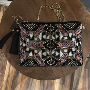 Mönstrad kuvertväska från Pipol's Bazaar - Snygg kuvertväska med ett färgglatt geometriskt mönster i svart, lila, rosa och vitt. Väskan har en elegant guldkedja och en dekorativ tofs. Perfekt för att ge din outfit en unik touch.