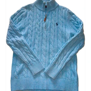 Ljusblå stickad tröja från Polo Ralph Lauren - Säljer en ljusblå stickad tröja från Polo Ralph Lauren med dragkedja och kabelstickat mönster. Tröjan har en broderad logotyp på bröstet och långa ärmar. Perfekt för kyliga dagar!
