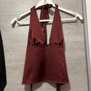 Vinröd halterneck linnetopp i satin - Säljer en vinröd halterneck linnetopp i glansigt satinliknande material. Toppen har djup v-ringning, knytning i nacken och baktill för en snygg passform. Perfekt för dig som vill sticka ut med en elegant och trendig look.