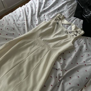 Elegant vit klänning med detaljer - Supersnygg vit långklänning med ärmlös design och vackra paljettbroderier vid halsringningen. Klänningen har en klassisk, figurnära passform upptill och ett utsvängt fall. Perfekt för dig som vill sticka ut med en stilren look.