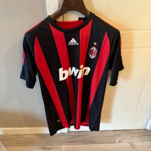 AC Milan Adidas fotbollströja röd/svart - Säljer en klassisk AC Milan fotbollströja från Adidas med röda och svarta vertikala ränder, klubbmärke och broderad stjärna på bröstet. Tröjan har vit Bwin-logga, korta ärmar och tre röda ränder på axlarna. Materialet är lätt och andas, perfekt för match eller träning. Helt ny, har andra tröjor om denna inte intressera dig!
