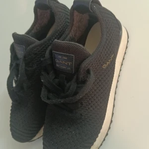 Svarta sneakers från Gant, strl 37 - Svarta sneakers från Gant med stickad ovandel och klassisk snörning. Snygg vit sula med svart yttersula och diskret Gant-logga på sidan och plösen. Perfekta för en clean och sportig look. Lätta och bekväma med modern design.