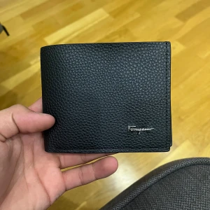 Ferragamo plånbok - Stilren svart plånbok från Ferragamo i exklusivt präglat skinn. Plånboken har flera kortfack och ett fönster för ID eller körkort. Perfekt storlek för fickan och diskret logga i silver på framsidan.