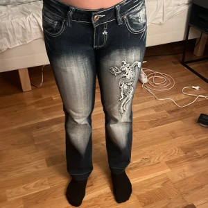 Blue Queen jeans med drakbrodyr - Snygga jeans från Blue Queen i mörkblå tvätt med slitningar och gråvita partier. Byxorna har broderade drakar på ben och bakfickor, klassisk femficksmodell och raka ben. Perfekta för dig som vill sticka ut med coola detaljer och streetkänsla. Jag sprättade upp dem ner till för de var något korta. Använt väldigt lite, säljer för att dem inte kommer till användning.