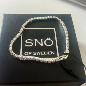 Silverfärgat armband från SNÖ of Sweden - Stilrent armband från SNÖ of Sweden i silverfärg med fyrkantiga länkar och gnistrande stenar. Klassisk och elegant design som ger en lyxig känsla. Perfekt detalj för att lyfta din outfit.