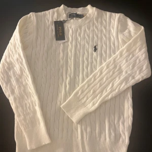 Vit kabelstickad tröja  - Vit kabelstickad tröja med klassisk rund halsringning och broderad logga på bröstet. Tröjan har lång ärm och ribbade muddar för en snygg passform. Perfekt för dig som gillar stilren och tidlös design.OBS INTE ÄKTA