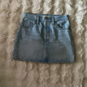 Ljusblå jeanskjol från Levi's - Snygg ljusblå jeanskjol från Levi's med klassisk femficksdesign, bälteshällor och knappstängning framtill. Kjolen har raka linjer och tydliga sömmar, samt Levi's ikoniska läderpatch och röd etikett bak. Perfekt skick, har inga defekter!
