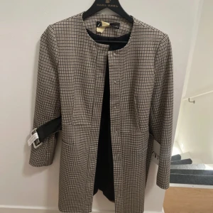 Rutig kappa med bälte i beige och svart - Snygg rutig kappa i beige och svart med klassiskt mönster och tillhörande bälte i midjan. Jackan har knappar framtill, rund halsringning och långa ärmar. Perfekt för dig som gillar en stilren och tidlös look. Fungerar också som storlek S!
