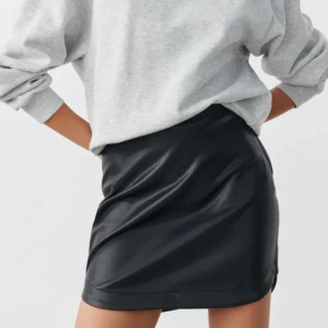 Svart skinnkjol - Snygg svart kort skinnkjol med rak passform och diskret dragkedja bak. Kjolen har en stilren look och passar perfekt till en oversized hoodie eller en cool t-shirt. Materialet är slätt och ger en edgy vibe.