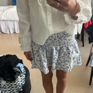 Vit och blå blommig kjol H&M - Supersöt kort kjol från H&M i vitt med blått blommönster. Kjolen har smock i midjan och volang nertill .