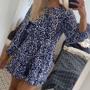 Blåvit kortklänning med mönster från Zara - Supersöt kortklänning i blått och vitt med unikt mönster och volangkant. Klänningen har långa ärmar med resår, markerad midja och snygg snörning vid halsen. Perfekt för dig som gillar boho-vibes och vill sticka ut lite extra.