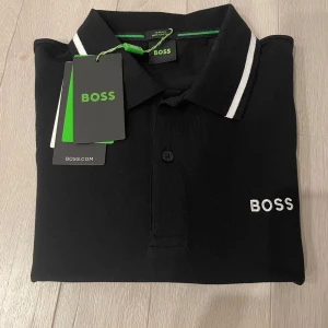 Svart pikétröja från BOSS -  Selling this brand new with tags BOSS polo shirt in sleek black. Size M (Men's). A timeless designer piece with minimalist white logo on the chest and sporty stripe details on the collar and sleeves. Made from high-quality cotton with a soft stretch feel - comfy, breathable, and built to last. Sharp fit that works for both casual and semi-formal looks. Style Tip: Pair with tailored shorts, jeans, or chinos. Perfect for date night, dinner, or casual Fridays. It's giving clean
