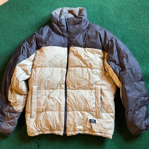 Beige och svart dunjacka Helly Hansen - Säljer en beige och svart dunjacka från Helly Hansen i storlek M. Jackan har hög krage, två stora fickor framtill och dragkedja. Perfekt för kalla dagar med sin quiltade design och sportiga look. Klassisk logga nertill på framsidan.