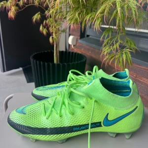 Nike Phantom GT2 fotbollsskor neon - Säljer ett par Nike Phantom GT2 fotbollsskor i neon-grönt med blå detaljer och svart Nike-logga. Skorna har snörning, stickad strumpkrage och mönstrad ovandel för extra grepp. Perfekta för dig som vill sticka ut på planen.