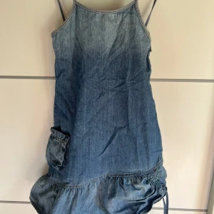 Jeansklänning med volang och ficka - Snygg jeansklänning från La Girls i 100% bomull. Modellen är kort med smala axelband, volang nertill och en cool ficka på sidan. Perfekt för dig som gillar avslappnad och trendig stil. Dragsko i nederkant för extra detalj.