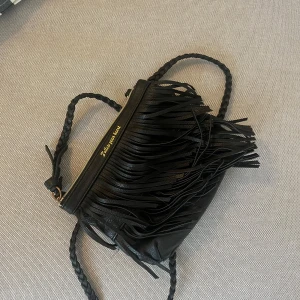 Svart axelväska med fransar - Svart axelväska i skinnimitation med flätat axelband och långa fransar framtill. Väskan har guldfärgad text 'Follow your heart' vid dragkedjan och en smidig storlek som passar till det mesta. Perfekt för dig som gillar detaljer och vill sticka ut lite extra.