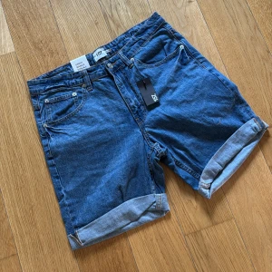 Blå jeansshorts från 157, midwaist - Klassiska blå jeansshorts från 157 med upprullade benslut och femficksdesign. Shortsen har normal passform och är tillverkade i slitstarkt denimtyg. Perfekta för sommardagar och enkel att matcha med allt. Snygga detaljer med synliga sömmar och bälteshällor.