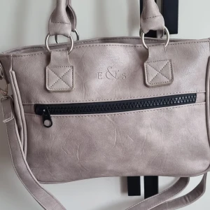 Beige axelväska från E&S med dragkedja - Snygg beige axelväska från E&S i syntetmaterial med svarta dragkedjedetaljer och silvriga ringar. Väskan har ett ytterfack med dragkedja och ett blommigt foder på insidan. Perfekt storlek för vardagens essentials och har både handtag och axelrem.