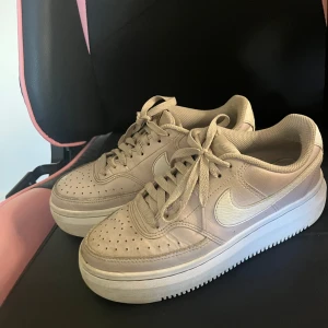 Beige Nike Air Force 1 sneakers - Snygga beige Nike Air Force 1 sneakers med vit sula och klassisk vit swoosh på sidorna. Skorna har snörning, perforerad tå och är tillverkade i skinn med meshfoder för extra komfort. Perfekta för dig som gillar stilrena och tidlösa sneakers.