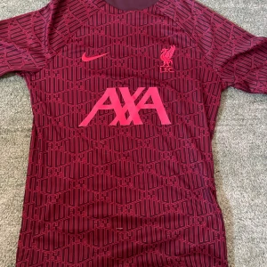 Liverpool tränings-tshirt Nike vinröd - Liverpool FC tränings-tshirt från Nike i vinröd med grafiskt mönster och rosa detaljer. AXA-logga på bröstet, LFC-märke och Nike Swoosh. Tillverkad i Dri-FIT-material som andas. Kortärmad och slim fit, perfekt för fotbollsträning.