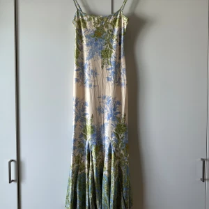 Lång blommig klänning med smala axelband - Superfin långklänning med smala axelband och blommigt mönster i blått och grönt på en ljus bas. Klänningen har en figurnära passform upptill och faller snyggt nedtill. Perfekt för sommarens alla tillfällen.