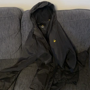 Svart vindjacka från Lyle & Scott - Svart vindjacka från Lyle & Scott med huva och klassisk gul logga på bröstet. Jackan har dragkedja framtill och två fickor, bra skick