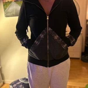 Svart hoodiejacka med snake-detaljer - Svart hoodiejacka från Elly Pistol med dragkedja och coola ormmönstrade detaljer på fickor och ärmslut. Jackan har snörning i huvan och är i mjukt material. Ena snöret har tappat metallpluppen som satt på 