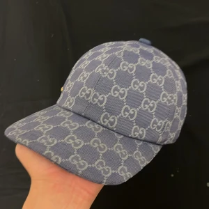 Blå Gucci keps - Snygg blå keps från Gucci med klassiskt GG-monogram i ljusare blått över hela kepsen. Kepsen har böjd skärm och justerbar baksida. Perfekt för dig som vill sticka ut med en lyxig accessoar.