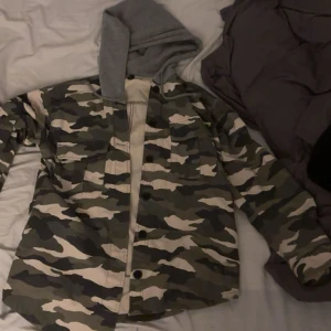 Camouflage overshirt med grå luva - Säljer en camouflagefärgad overshirt från H&M Divided i storlek 38. Jackan har grå huva i sweatshirtmaterial, knäppning framtill och två bröstfickor. Perfekt för dig som gillar streetstyle och vill sticka ut lite extra. 