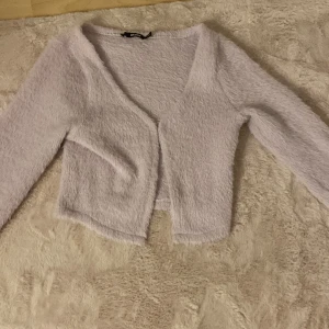 Ljuslila fluffig kofta från BikBok - Supermjuk och fluffig ljuslila kofta från BikBok i croppad modell. Koftan har lång ärm, v-ringad hals och är tillverkad i ett stickat material som känns riktigt cozy. Perfekt att slänga på sig för en chill look.
