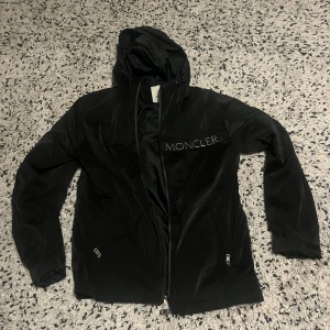 Svart vindjacka från Moncler med huva - Svart vindjacka från Moncler med huva och dragkedja framtill. Jackan har Moncler-logga på bröstet, två fickor med dragkedja och en lätt glansig finish. Perfekt för dig som vill ha en clean och stilren look. Innerfoder med coolt serietryck.