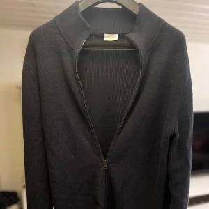 Mörkblå stickad kofta John Henric - Mörkblå stickad kofta från John Henric med hel dragkedja och hög krage. Klassisk och stilren modell i mjukt material, perfekt för lager-på-lager. Långärmad och ribbad struktur som ger en snygg och enkel look.