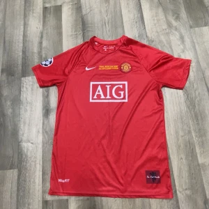 Manchester United Ronaldo 7 tröja - Klassisk röd Manchester United fotbollströja från Nike med AIG som sponsor, Champions League-märke på ärmen och tryck 'Ronaldo 7' på ryggen. Tröjan har korta ärmar och är tillverkad i lätt, ventilerande material perfekt för match eller träning.