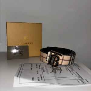 Burberry rutig skärp med logga - Snyggt Burberry-bälte med klassiskt rutigt mönster i beige, svart och rött. Bältet har en stor metallspänne med Burberrys ikoniska TB-logga i svart och guld. Perfekt accessoar för att lyfta din outfit med lyxig känsla.
