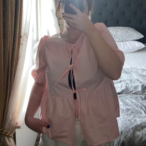 Ljusrosa blus med puffärm och knyt - Supersöt ljusrosa blus med puffiga korta ärmar och tre knytband framtill. Blusen har en lös passform och volang nedtill som ger en romantisk vibe. Perfekt för dig som gillar gulliga detaljer och en feminin look.