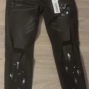 Svarta jeans med vit färgdetalj - Säljer ett par svarta jeans med coola vita färgstänk och slitningar för en edgy look. Jeansen har smal passform och klassisk femficksdesign. Perfekta för dig som vill sticka ut med en unik streetstyle.