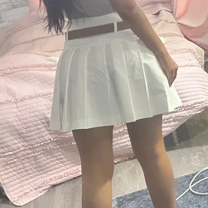 Oanvänd vit plisserad kortkjol med medföljande brunt bälte  - Superfin vit plisserad kortkjol med hög midja och brunt bälte i midjan. Kjolen har klassiska veck och ger en fräsch och sportig vibe. Perfekt för dig som gillar stilrena och trendiga outfits.