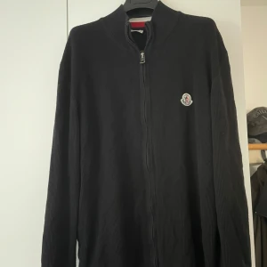 Svart stickad kofta från Moncler - Svart stickad kofta från Moncler med dragkedja framtill och klassisk logga på bröstet. Långa ärmar med ribbade muddar och snygg detalj med röd, vit och blå rand vid ärmslutet.