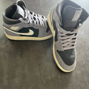 Nike Air Jordan 1 grå och svarta - Snygga Nike Air Jordan 1 sneakers i grått och svart med vita detaljer och klassisk vinge-logga på sidan. Skorna har högt skaft, snörning och är tillverkade i skinn. Perfekta för dig som vill ha en ikonisk och streetig look.
