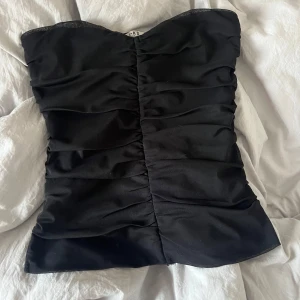 Svart tubtopp från NA-KD XS - Säljer en svart tubtopp från NA-KD i storlek XS. Toppen har snygga rynkade detaljer framtill och baktill som ger en cool och trendig look. Gjord i ett mjukt och stretchigt material som sitter skönt och formar sig efter kroppen. Perfekt för att lyfta vilken outfit som helst.
