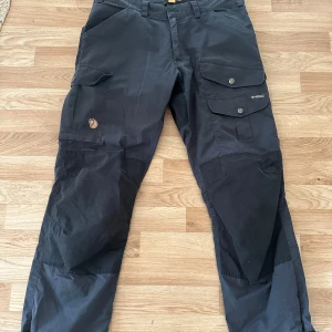 Svarta cargopants från Fjällräven EU 50 - Svarta cargopants från Fjällräven i slitstarkt G-1000-material. Byxorna har flera stora fickor med knappar på benen, förstärkta knän och justerbara benslut. Perfekta för dig som gillar outdoor och vill ha både funktion och stil.