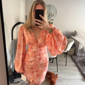 Adoore - Superfin kort klänning i ljusa orange och rosa toner med blommigt mönster. Djup v-ringning, markerad midja och knappar framtill. Stora puffärmar ger en trendig look. Perfekt för sommarens alla tillfällen! HELT oanvänd med lapp, pris gäller! Pris i butik 1500